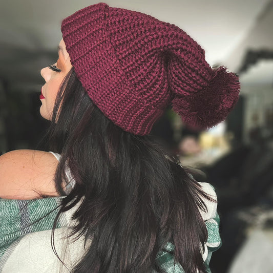 A woman with long dark hair faces sideways, wearing the Chunky Ribbed Pom Beanie in maroon. She pairs it with a white top and a green plaid wrap. The softly blurred backdrop adds warmth to this stylish acrylic accessory.