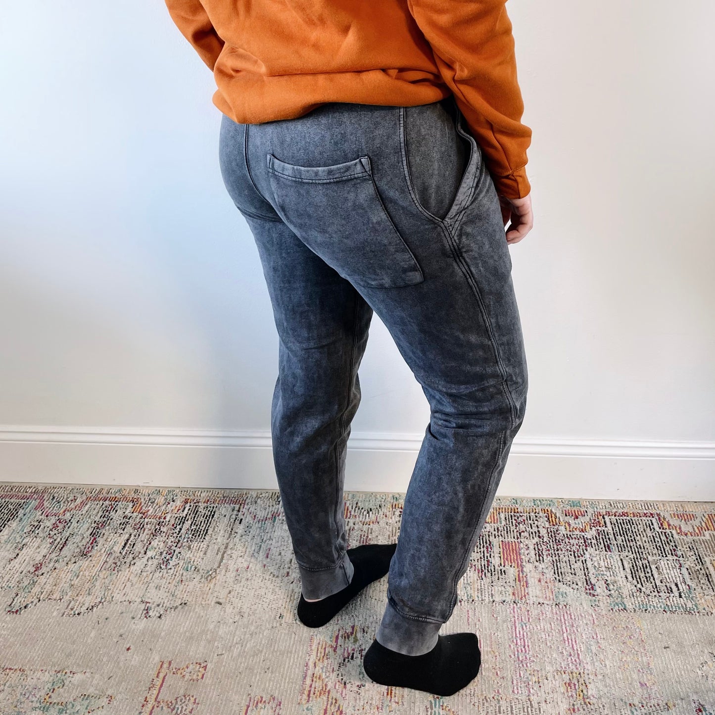 Person stands indoors on a patterned rug facing away from the camera, wearing Mama Sporty • Acid Wash Joggers, an orange long-sleeve shirt, and black socks against a plain white wall.
