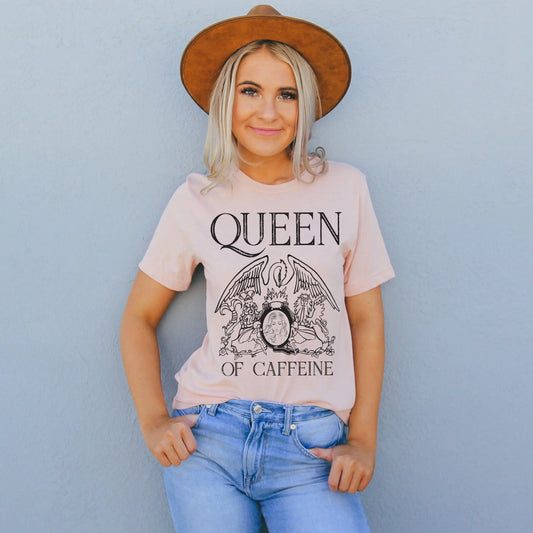 A smiling woman wearing a Queen • Peach Tee and a brown hat stands against a gray wall with one hand in her jean pocket.
