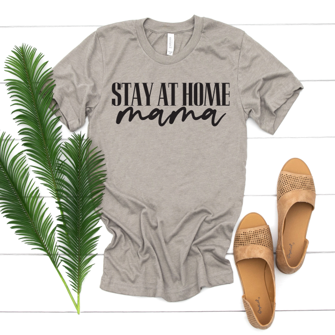 The Stay at Home Mama • Tee by Bella Canvas in gray heather stone features “STAY AT HOME mama” in black. Shown on a white wood background beside tan sandals and green palm leaves—perfect for dedicated stay-at-home moms.
