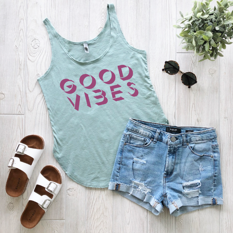 Flat lay of the Good Vibes • Sage Tank with pink lettering, paired with distressed denim shorts, white sandals, black sunglasses, and a small green plant on a white wooden surface.