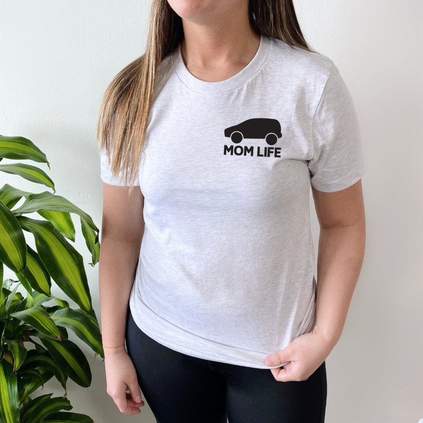 A woman with long brown hair stands indoors next to a green leafy plant, wearing the Mini-Vans • Tee Shirt—a light gray Bella/Canvas top featuring a VANS-inspired black minivan graphic and MOM LIFE printed on the chest.