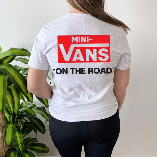A person in a white Mini-Vans • Tee Shirt with a red and white MINI-VANS graphic and black ON THE ROAD text stands next to a green leafy plant—a perfect VANS-inspired look for everyday mom life.