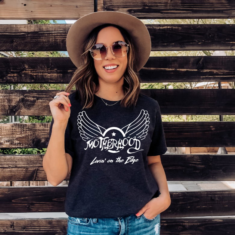 A smiling woman in sunglasses, a wide-brim hat, and the Aerosmith Mama Tee • Charcoal stands in front of a wooden fence on a sunny day.