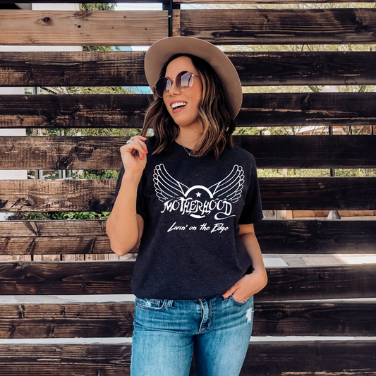 A woman in sunglasses, a wide-brim hat, and the charcoal Aerosmith Mama Tee with a unisex fit smiles by a wooden fence, one hand in her pocket and the other holding a lollipop.