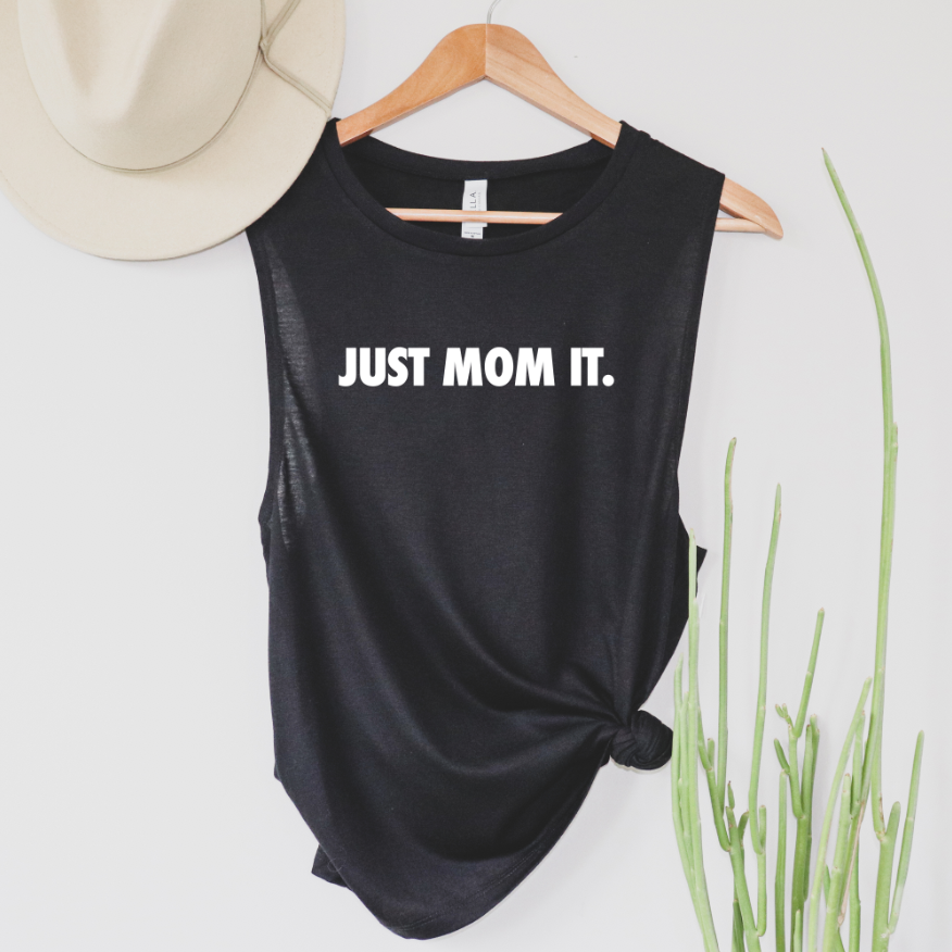 A black JUST MOM IT • Scoop Tank hangs on a wooden hanger, with a beige hat above and a green plant to the right, highlighting this chic womens flowy tank top.