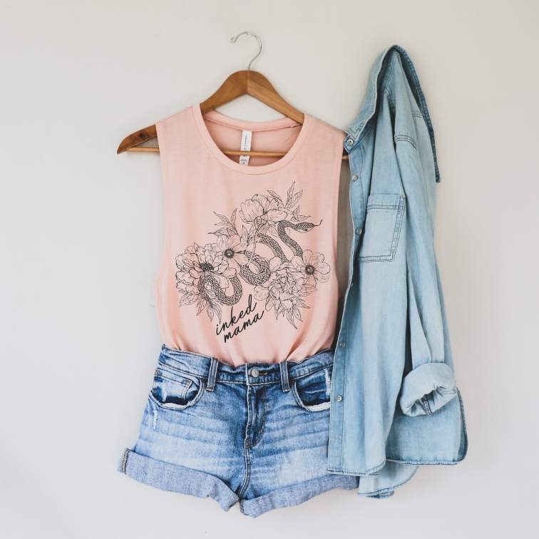 The Floral Snake • Scoop Tank, a light pink flowy women’s top with “mama” text and a floral snake print, is styled with high-waisted denim shorts and a light blue shirt on a wooden hanger against a white wall.