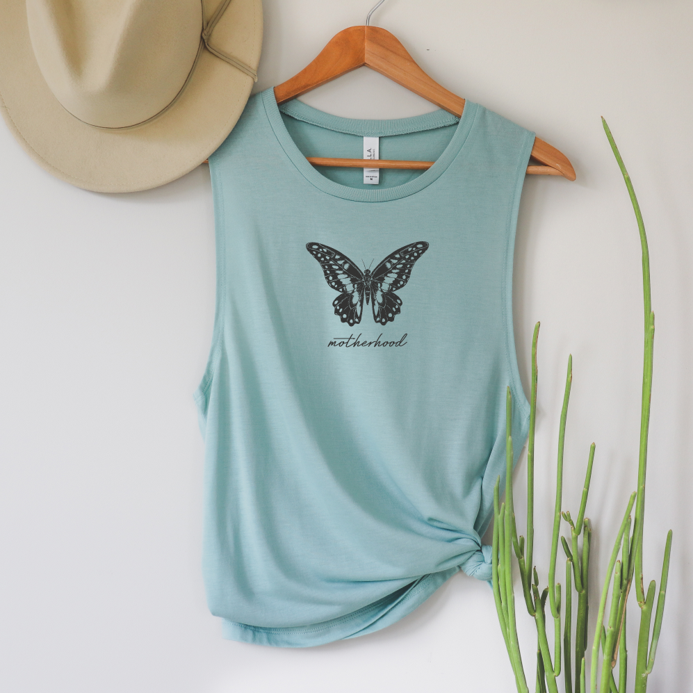 The Butterfly • Scoop Tank is a light blue womens relaxed-fit tank featuring a black butterfly and motherhood print. Displayed on a wooden hanger, its styled with a beige hat and tall green plant against a white wall.