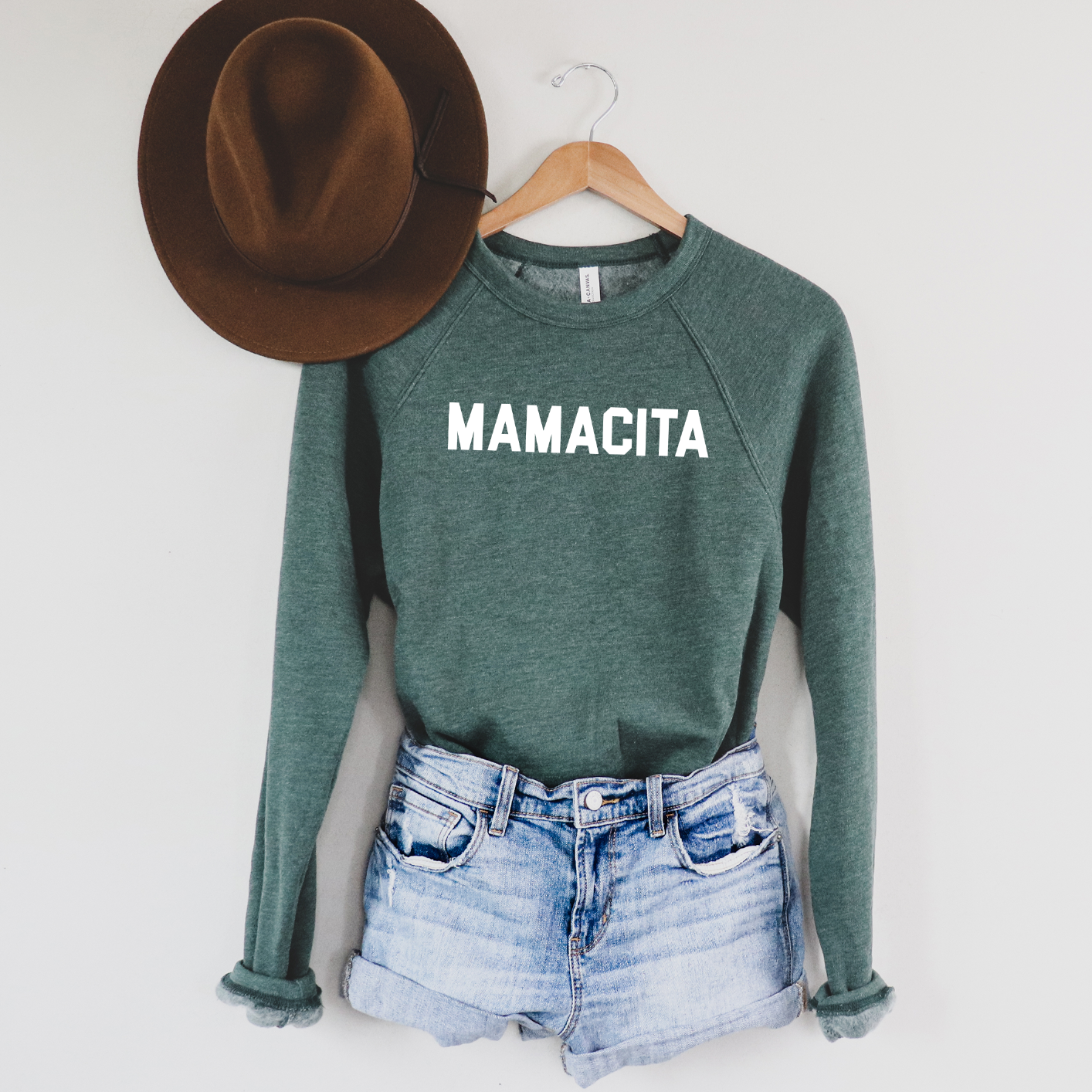The MAMACITA • Forest Pullover in green with a bold white MAMACITA print is displayed on a wooden hanger, paired with blue denim shorts and a brown wide-brimmed hat against a light-colored wall for a stylish unisex look.