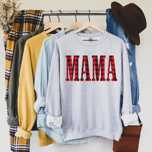 A rack of casual clothes, including a plaid shirt, denim shirt, and the MAMA • Plaid Print Pullover with bold red plaid letters. Nearby, a brown hat and purse with dried flowers add a cozy accent.
