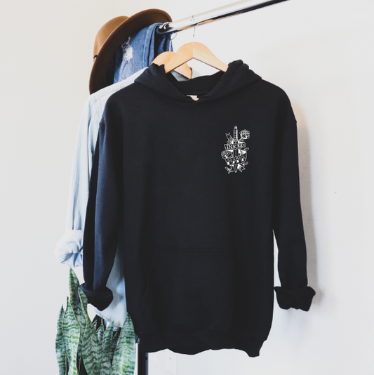 The Inked Mama • Black Hoodie, a black unisex fit with a small white graphic, hangs on a wooden hanger; behind it are a blue button-up shirt and a brown hat on the rack, with green plants below.