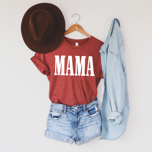 The MAMA • Clay Tee, a unisex fit red Bella Canvas shirt with bold white MAMA block print, is styled with denim shorts, a light blue chambray shirt, and a brown hat on a wooden hanger against a plain white wall.