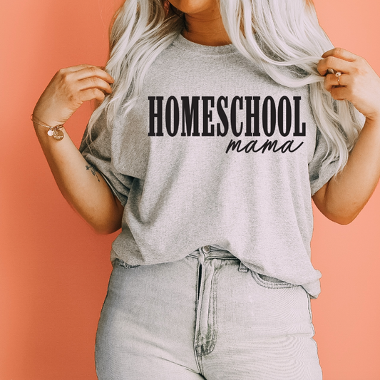 A woman with long blonde hair wears the New Homeschool Mama • Gray Tee from Bella Canvas, featuring bold black HOMESCHOOL mama text. She stands against a peach background, holding her shirt collar.
