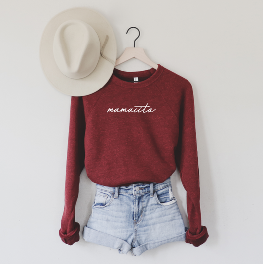 A wide-brimmed hat hangs on a white wall above a hanger with the mamacita • Cardinal Pullover in a unisex fit, paired with light denim shorts.