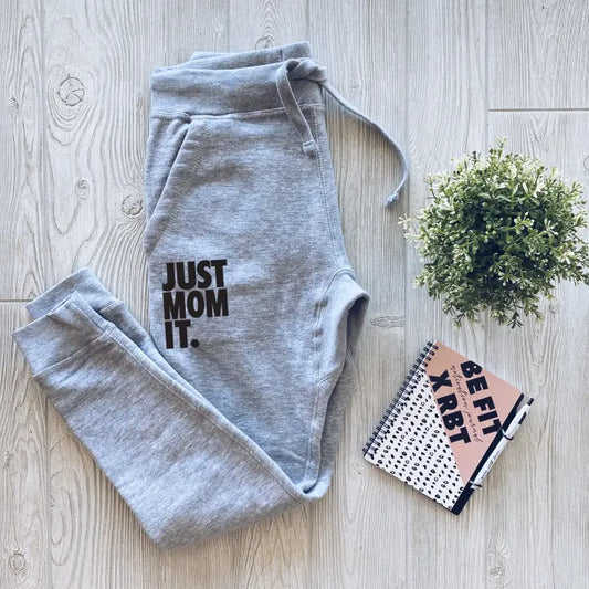 Just Mom It • Gray Joggers: Unisex fleece joggers in gray with JUST MOM IT. printed on the left leg, styled on a light wood floor next to a green potted plant and spiral notebook—perfect cozy loungewear for comfort and style.