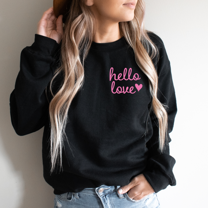 A woman with long, wavy blonde hair wears the Hello Love • Black Pullover featuring hello love and a pink heart, paired with light blue jeans. She holds a brown hat by her head. This classic unisex sweatshirt offers a comfy fit.