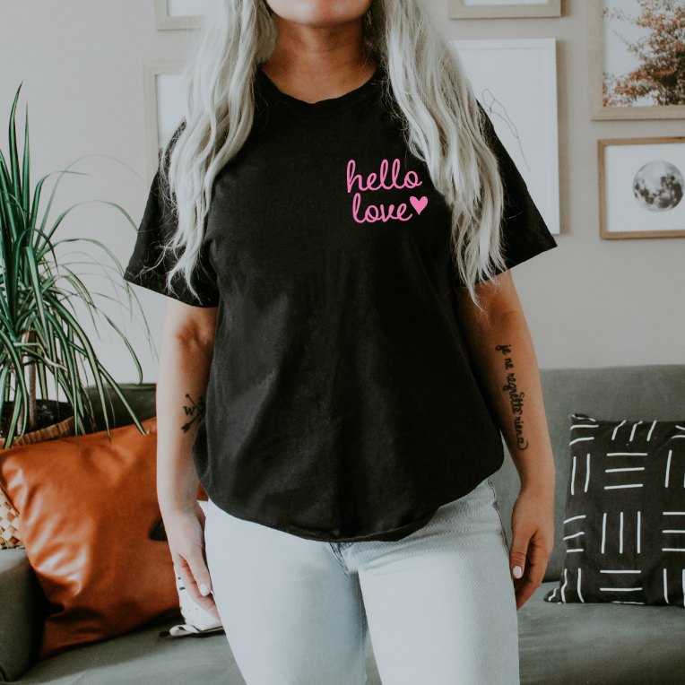 A person with long, light hair wears the Hello Love • Black Tee featuring “hello love” and a pink heart. They’re standing in a living room with a gray couch, orange pillow, and framed wall art in the background.