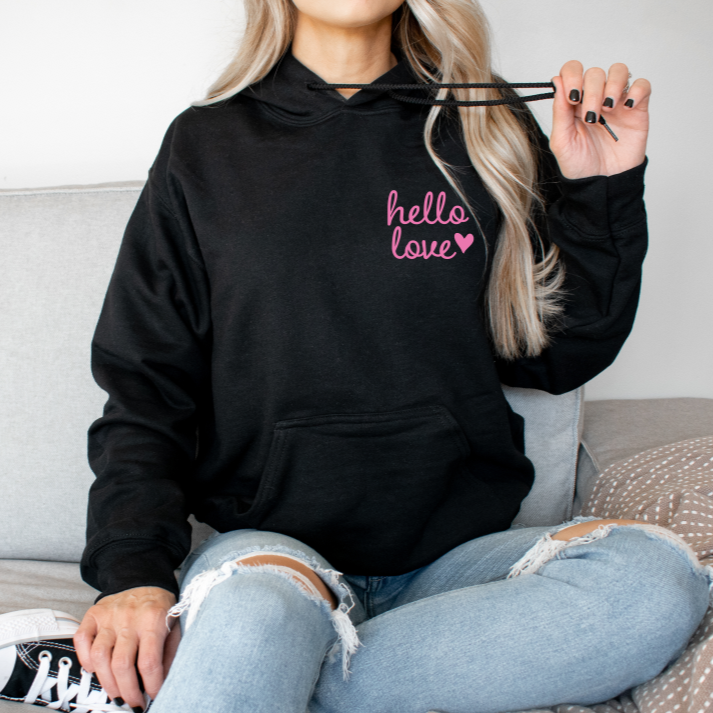 A person sits on a couch wearing the Love Print • Black Hoodie with hello love and a pink heart, paired with light blue ripped jeans, holding the hoodie strings in one hand.
