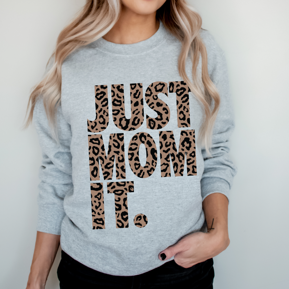 A woman with long, wavy blonde hair models the JUST MOM IT • Cheetah Pullover, a comfy unisex light gray sweatshirt with bold cheetah print lettering. She stands against a plain background, one hand tucked in her pocket.