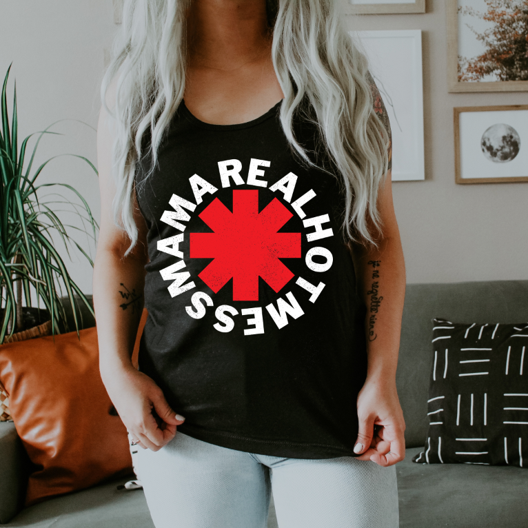 A person with long, wavy, light hair wears a black “Real Hot Mess Mama” unisex tank top in a modern living room featuring a couch, pillows, and framed wall art.