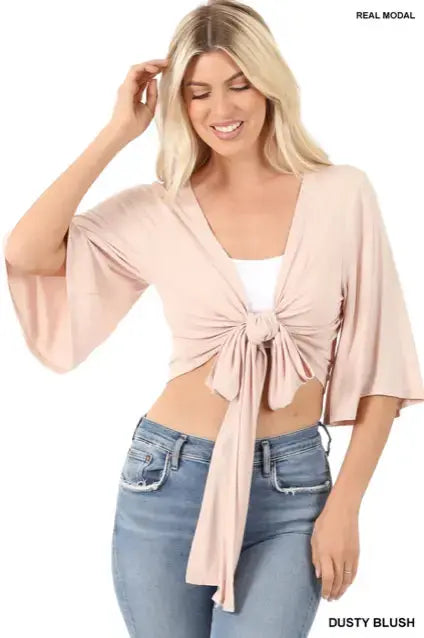 A woman wears the Jasmine Cardigan in dusty blush—a tie-front crop top with wide sleeves—over a white tank and blue jeans, smiling as she looks down and touches her hair. More colors available!.