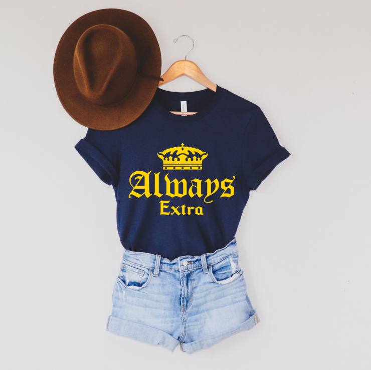The Always Extra • Navy Tee with a yellow crown graphic is styled with a brown hat and light blue denim shorts, all displayed on a hanger against a white background for a trendy unisex look.