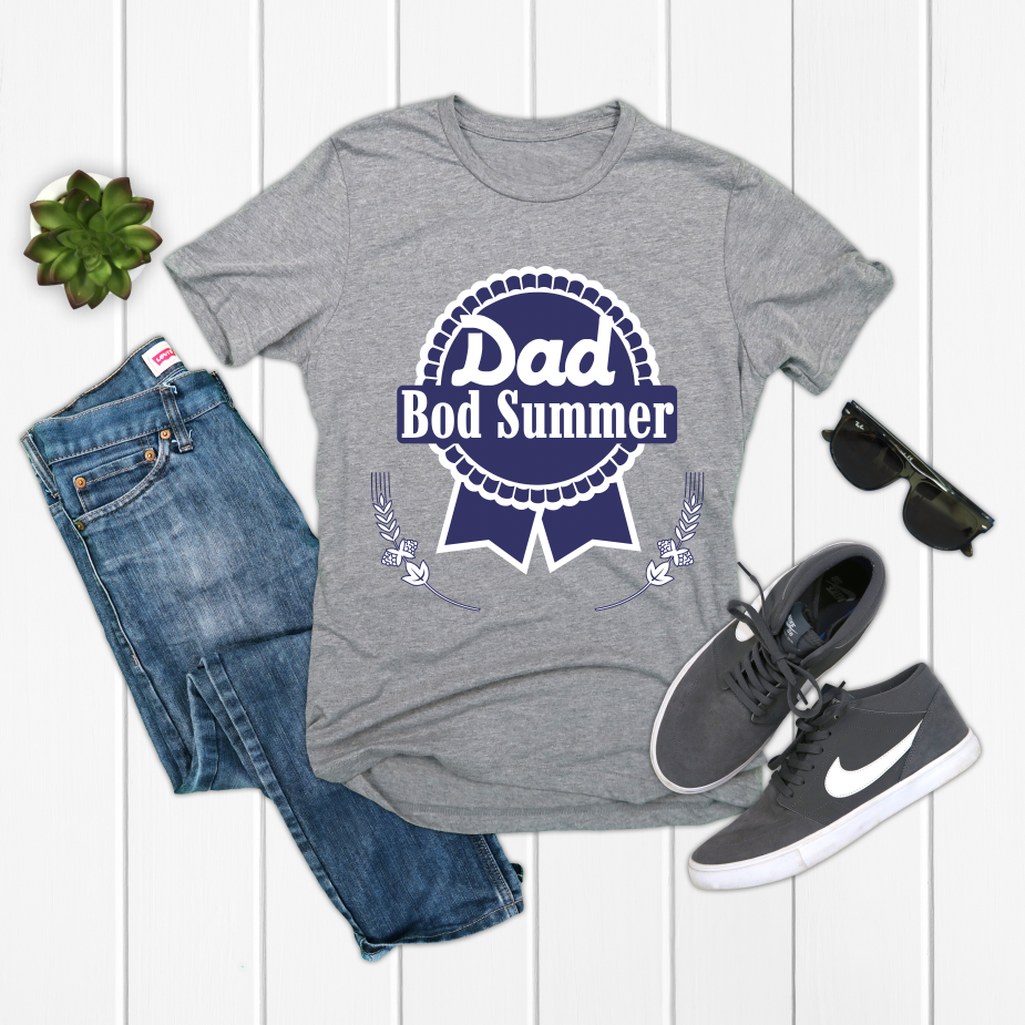 The Dad Bod Summer • Gray Tee features bold text on soft gray combed ring-spun cotton, styled with blue jeans, gray Nike sneakers, black sunglasses, and a green succulent on a white wooden surface.