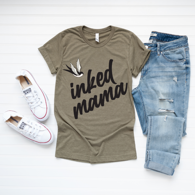 Flat lay of an Inked Mama • Olive Tee featuring a flying bird and inked mama text, styled with light blue ripped jeans and white sneakers on a white wooden background.