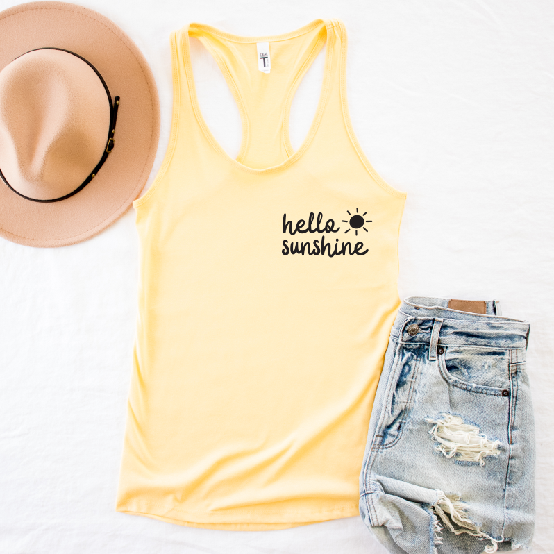 The Hello Sunshine • Yellow Tank is a soft, yellow racerback tank top featuring a hello sunshine design and sun graphic, made from combed ringspun cotton, displayed beside a beige hat and ripped denim shorts on a white surface.