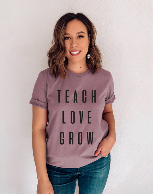 A woman with shoulder-length brown hair smiles at the camera, wearing the Teach. Love. Grow. • Orchid Tee Shirt and dark blue jeans, standing against a plain white wall.