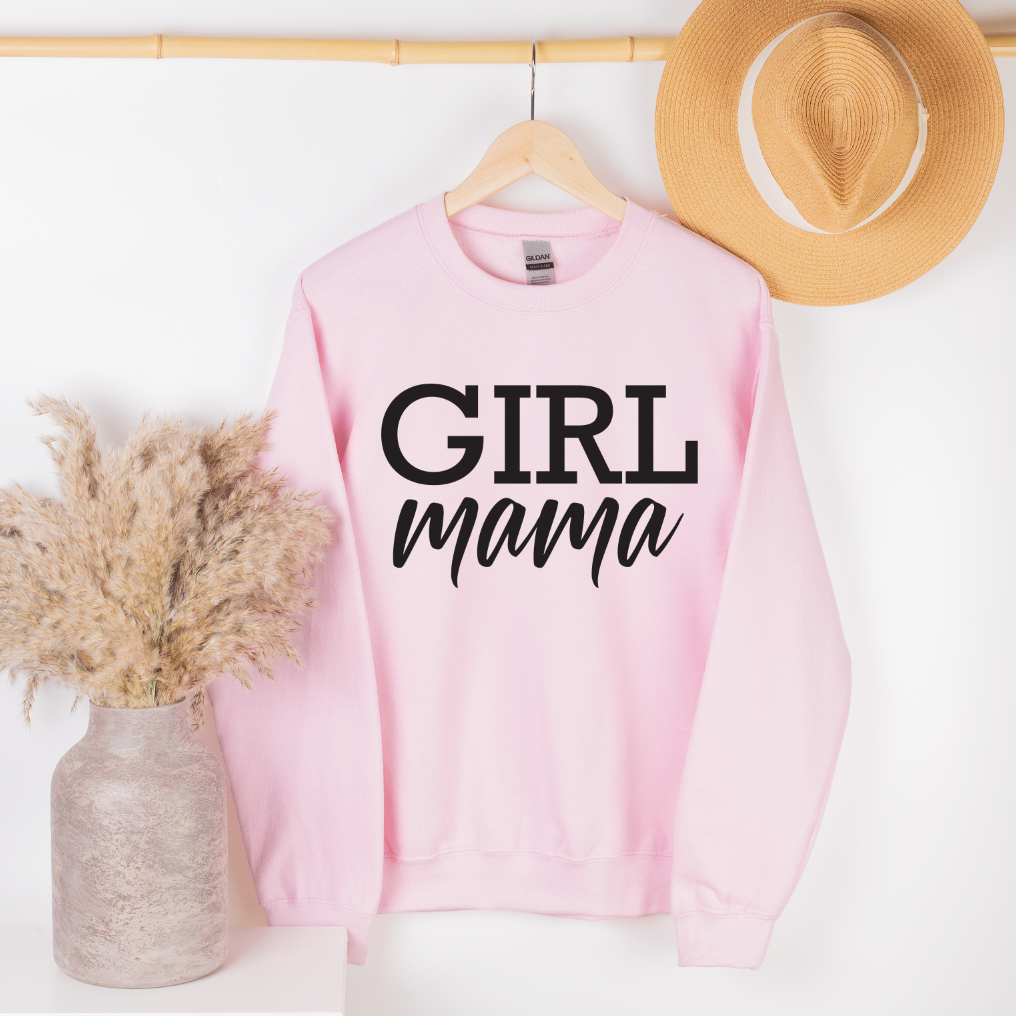 The Girl Mama • Pink Pullover, a light pink preshrunk cotton/polyester sweatshirt with “GIRL mama” in bold black letters, hangs on a rack beside a straw hat. Nearby, a white surface holds a vase with pampas grass.