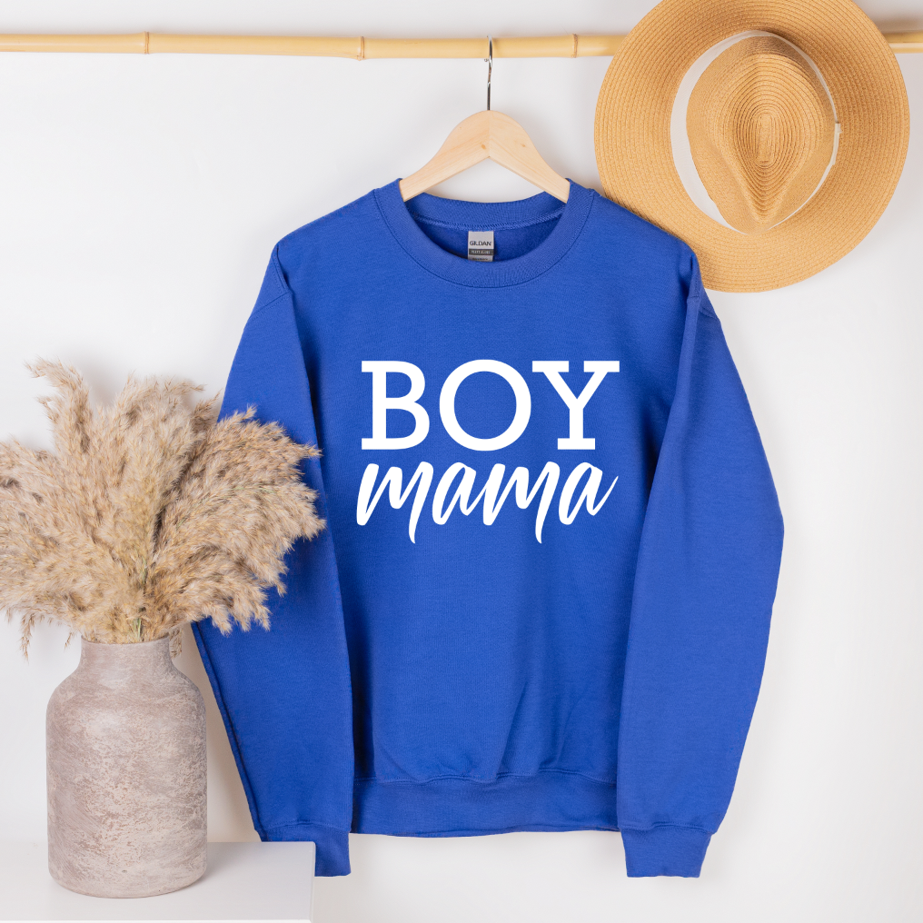 The Boy Mama • Royal Pullover, featuring boy mama in white on royal blue preshrunk cotton/polyester, hangs on a wooden hanger with a tan hat on the wall and pampas grass in a vase beside a white table.