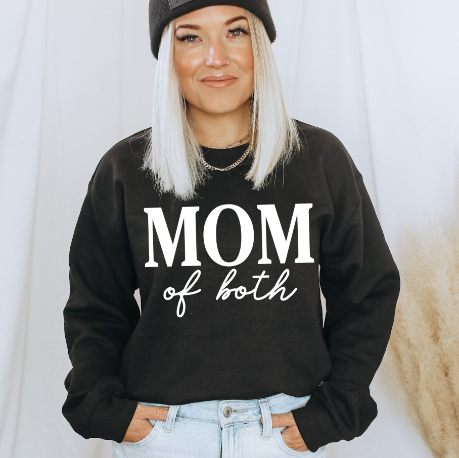 Woman with long blonde hair wears the Mom of both • Pullover, a black fleece unisex sweatshirt with white text, styled with a black beanie and hands in her pockets against a light background.