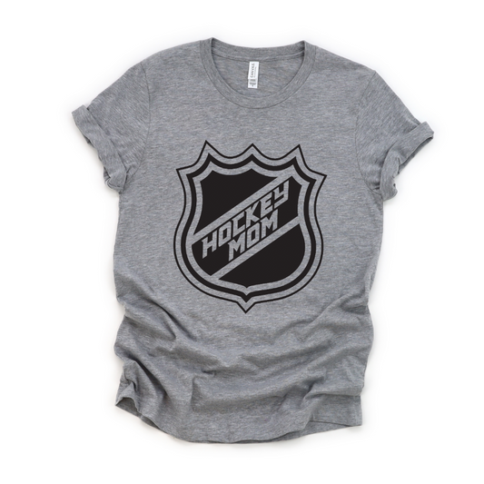 The Hockey Mom Tee is a heather gray unisex t-shirt with rolled sleeves, featuring a bold black hockey-style shield logo and HOCKEY MOM lettering—perfect for stylish and proud hockey moms.