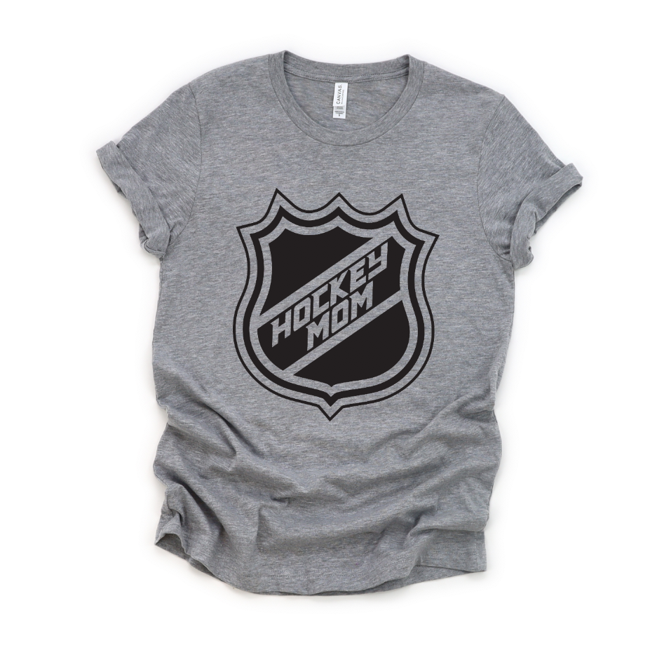 The Hockey Mom Tee is a heather gray unisex t-shirt with rolled sleeves, featuring a bold black hockey-style shield logo and HOCKEY MOM lettering—perfect for stylish and proud hockey moms.