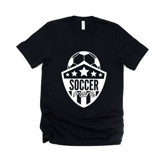 The Soccer Mom Tee is a black unisex shirt with a white soccer ball graphic above a stars-and-stripes shield and “Soccer Mom” printed in bold and script fonts.