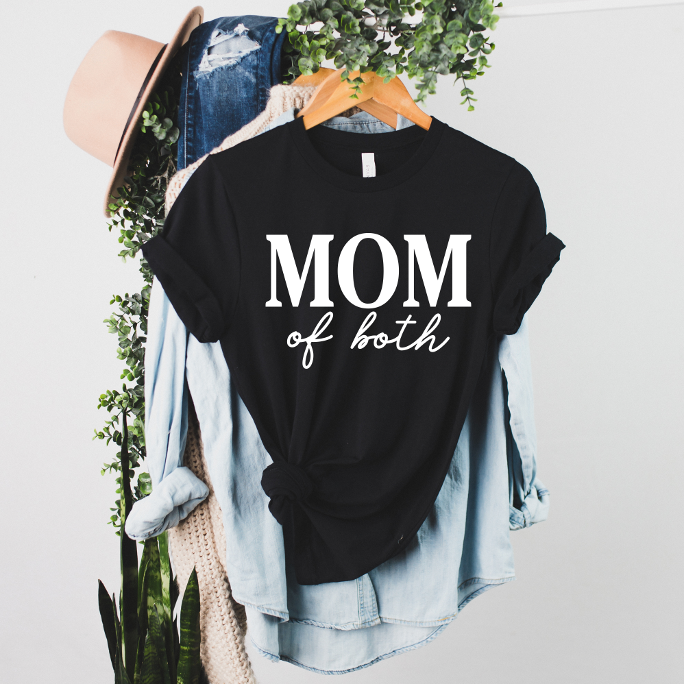 The Mom of both • Black Tee, a unisex Bella Canvas shirt with white MOM of both text, is shown on a hanger over a light denim shirt, surrounded by green plants, a beige hat, and casual clothes in the background.