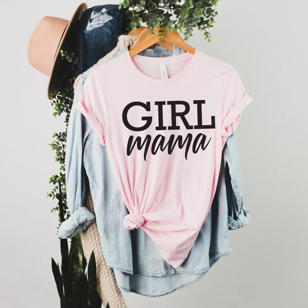 The Girl Mama • Pink Tee in light pink is styled knotted over a denim shirt on a rack with green plants, a beige sweater, jeans, and a tan hat—the ideal comfy mom tee for casual style.