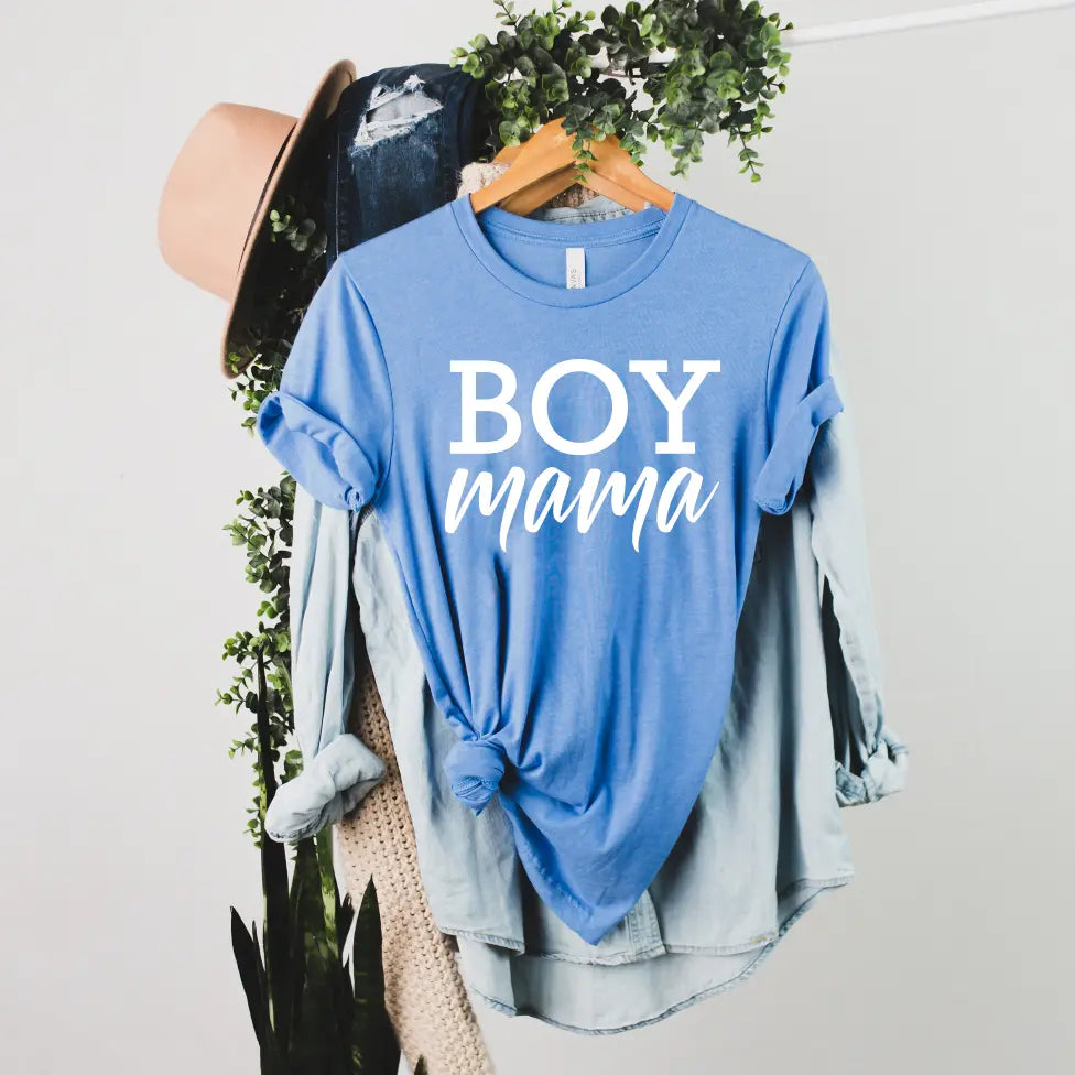 The Boy Mama • Royal Heather Tee is a comfy Bella Canvas blue shirt with BOY mama in white text, styled tied at the waist and layered over a denim shirt, shown on a hanger with plants, a hat, and jeans in the background.