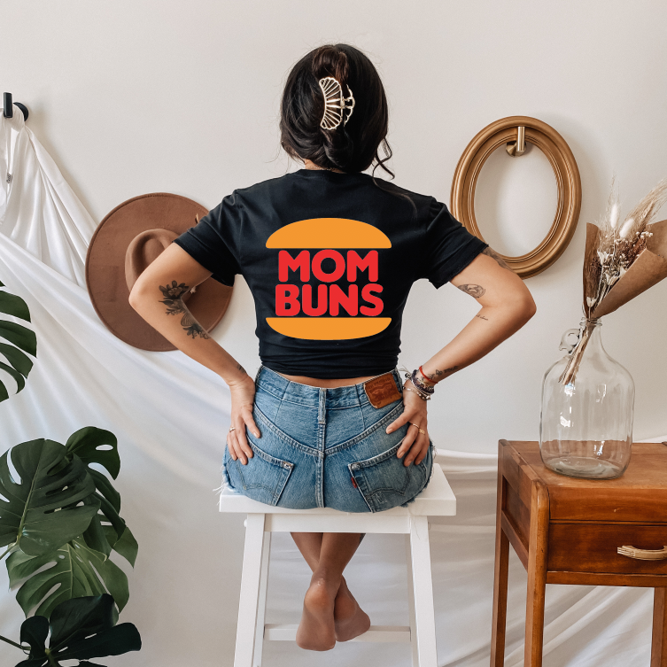 A woman with a hair bun sits on a white stool, facing away, wearing the MOM BUNS • Black Tee by Bella Canvas—100% cotton, featuring a burger graphic and MOM BUNS text. The room has neutral and natural accents.
