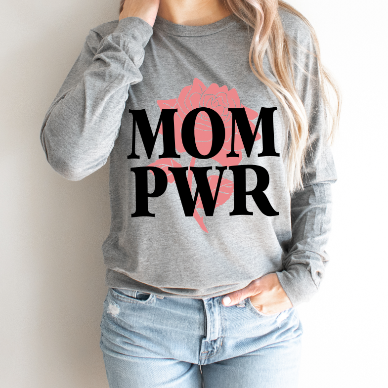 A person wears the MOM PWR • Long Sleeve—a grey shirt with bold black MOM PWR lettering over a pink rose graphic—celebrating moms superpower, paired with light blue jeans. Unisex sizing for a comfortable fit.