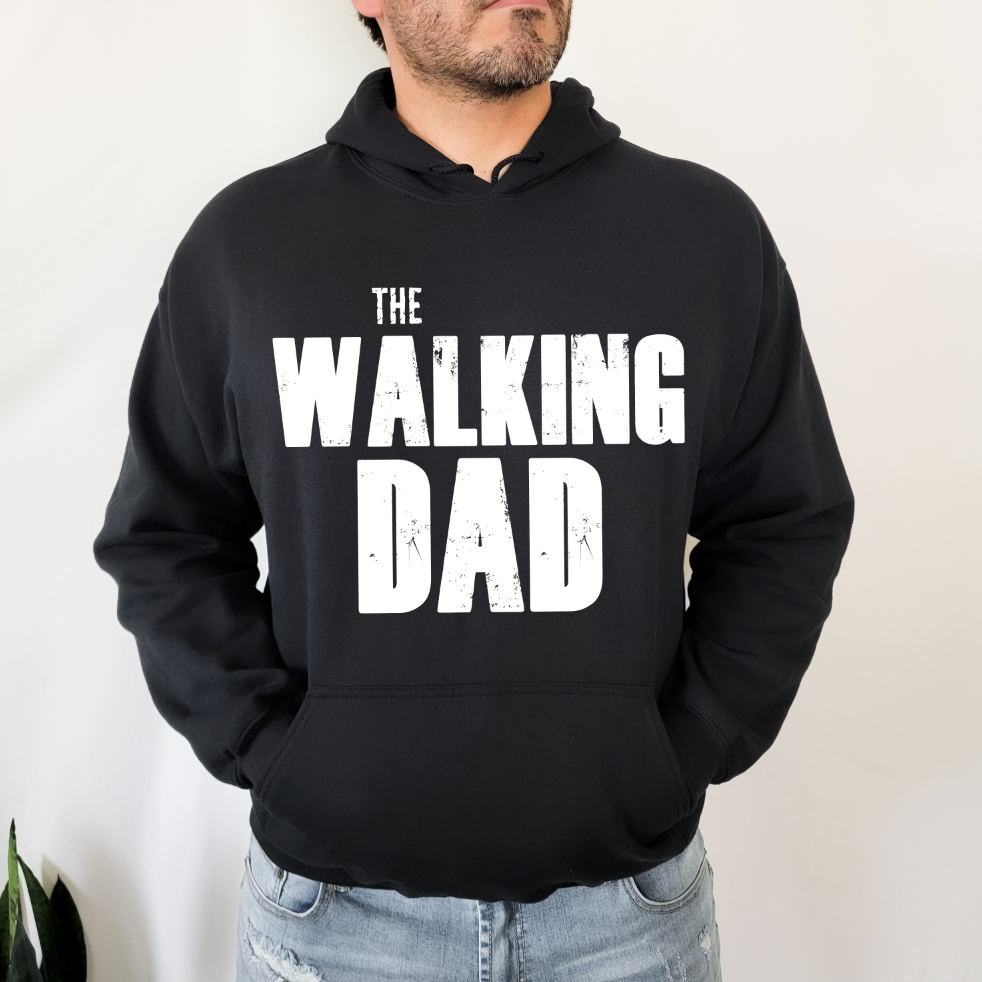 A man stands with hands in his pockets, wearing The Walking Dad • Black Hoodie—crafted from pill-resistant fleece and featuring bold white THE WALKING DAD lettering on the front.