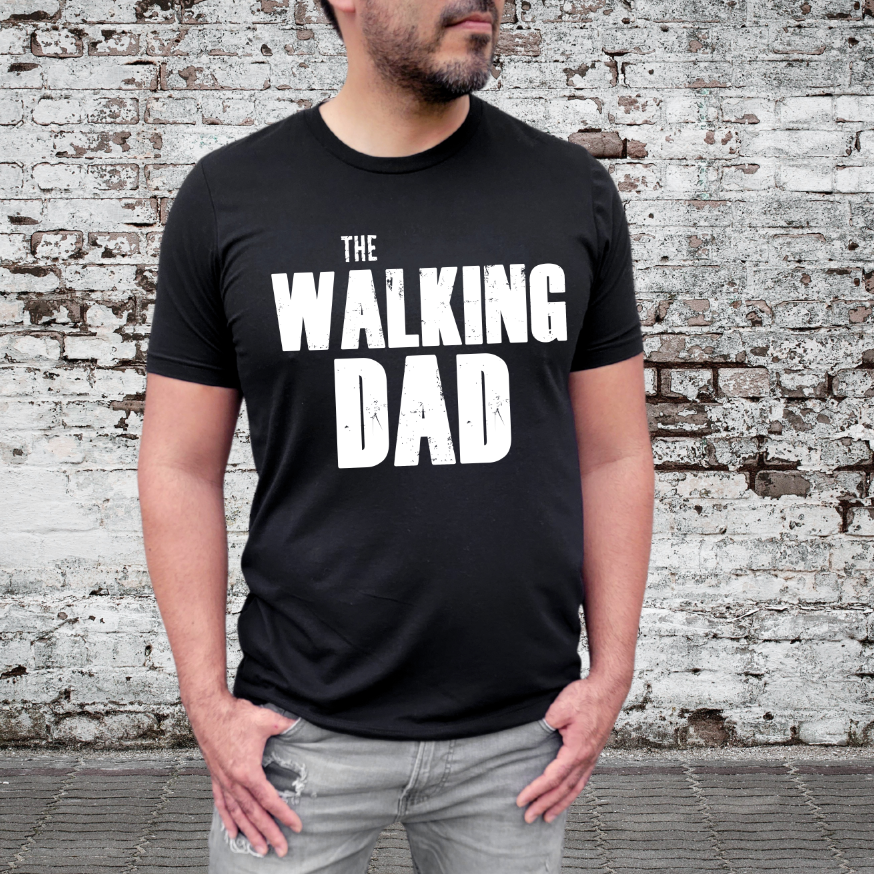 A man in ripped jeans and a black The Walking Dad • Adult Tee stands by a weathered brick wall, the bold white text making this unisex cotton shirt perfect for zombie dads everywhere.
