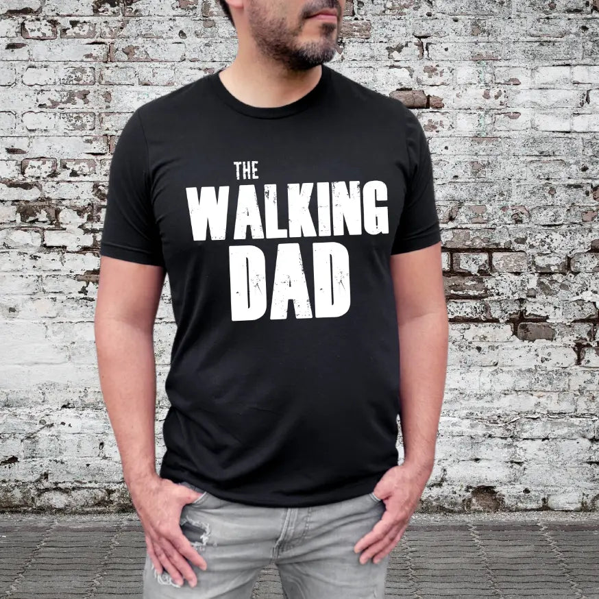 A man in ripped jeans and a black The Walking Dad • Adult Tee stands by a weathered brick wall, the bold white text making this unisex cotton shirt perfect for zombie dads everywhere.
