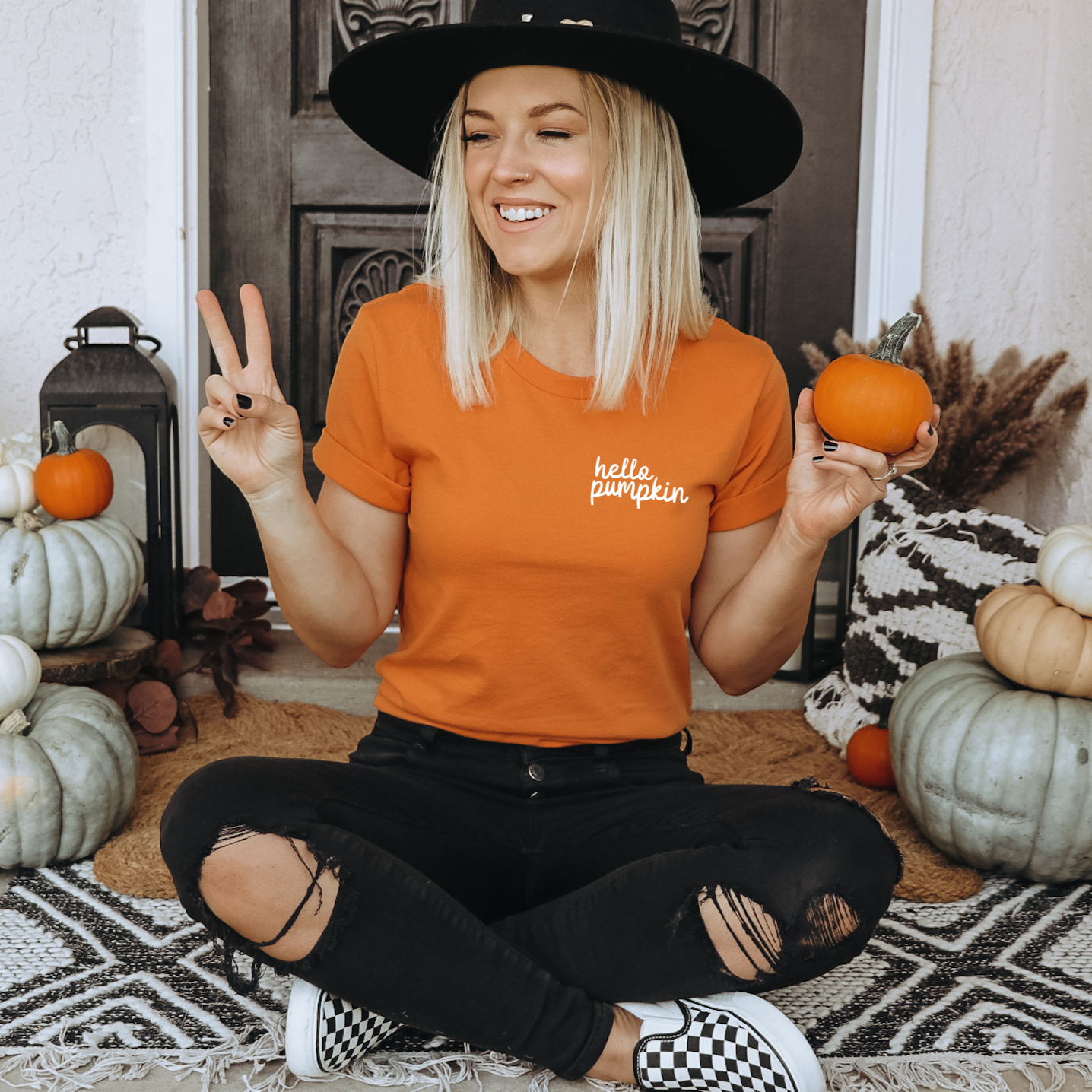 A smiling woman in a Hello Pumpkin • Orange Tee and black hat sits cross-legged on a porch with pumpkins, holding a small pumpkin in one hand and making a peace sign with the other.