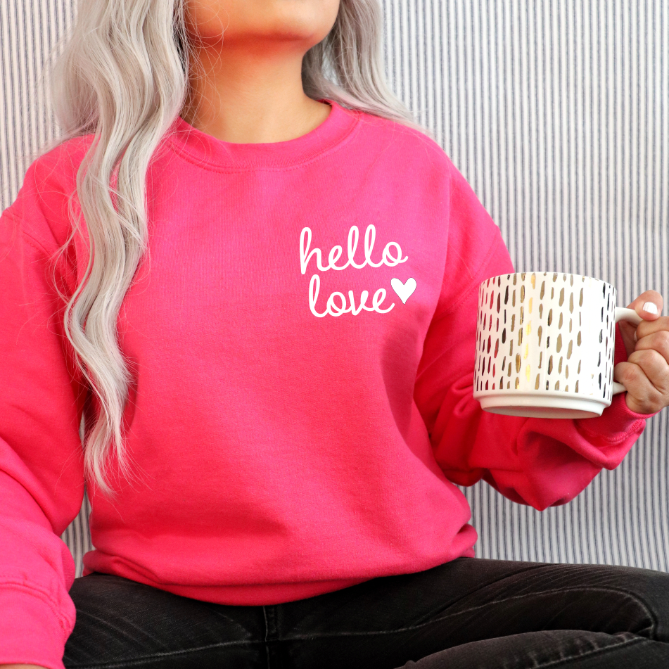 A person with long, wavy, light hair wears the hello love • Bright Pink Pullover and holds a large white mug with gold and black patterns against a striped background.