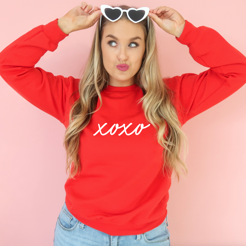 A woman wears the xoxo • Red Pullover, a unisex Valentines Day sweatshirt, with light blue jeans. She poses playfully against a pink background, holding white heart-shaped sunglasses above her head and making a pouty face.