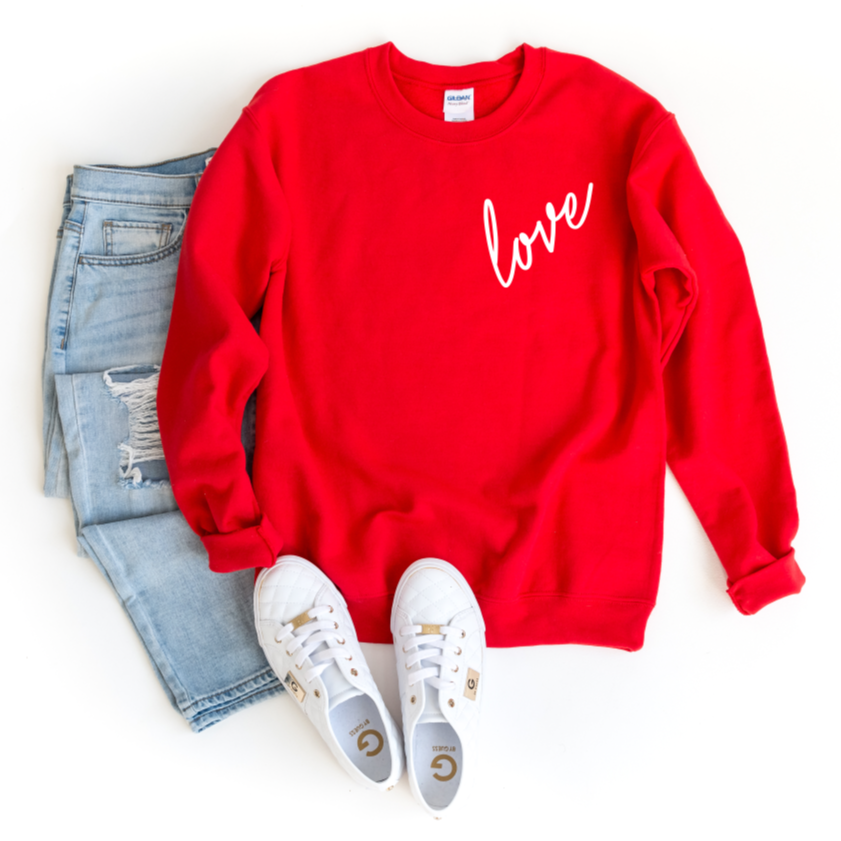 The Love • Red Pullover, a comfy unisex sweatshirt with “love” in white script, styled with light blue ripped jeans and white sneakers on a white background—perfect for any casual Valentine’s Day look.