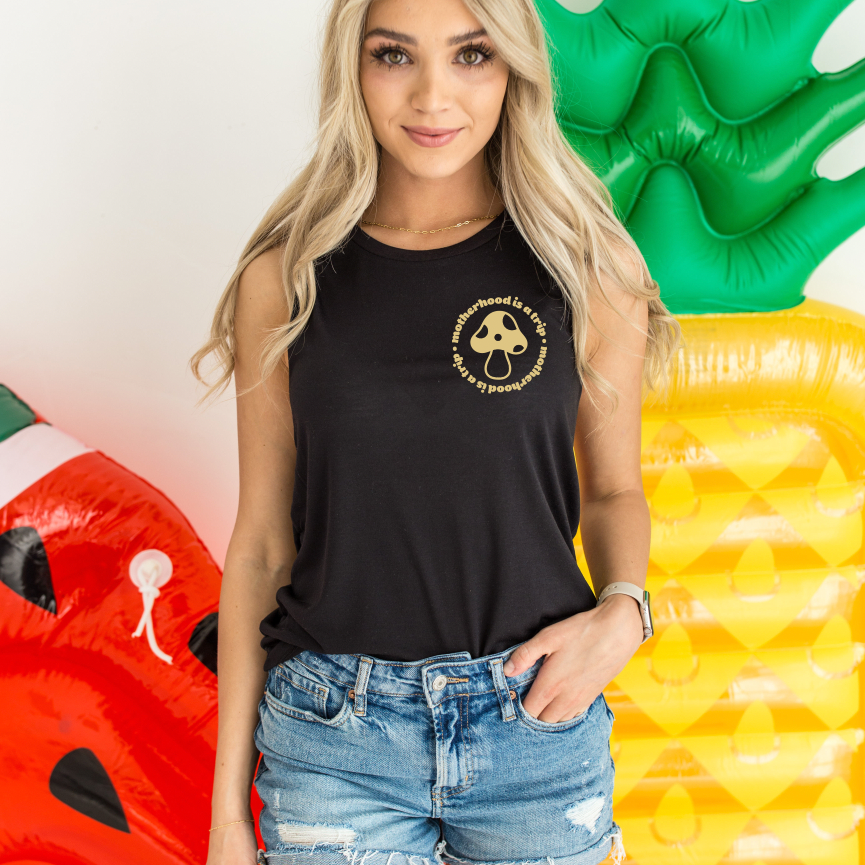 A woman with long blonde hair wears the Motherhood is a Trip • Scoop Tank, a relaxed fit black sleeveless top with mushroom and text print, paired with distressed denim shorts. She stands in front of inflatable watermelon and pineapple decorations.