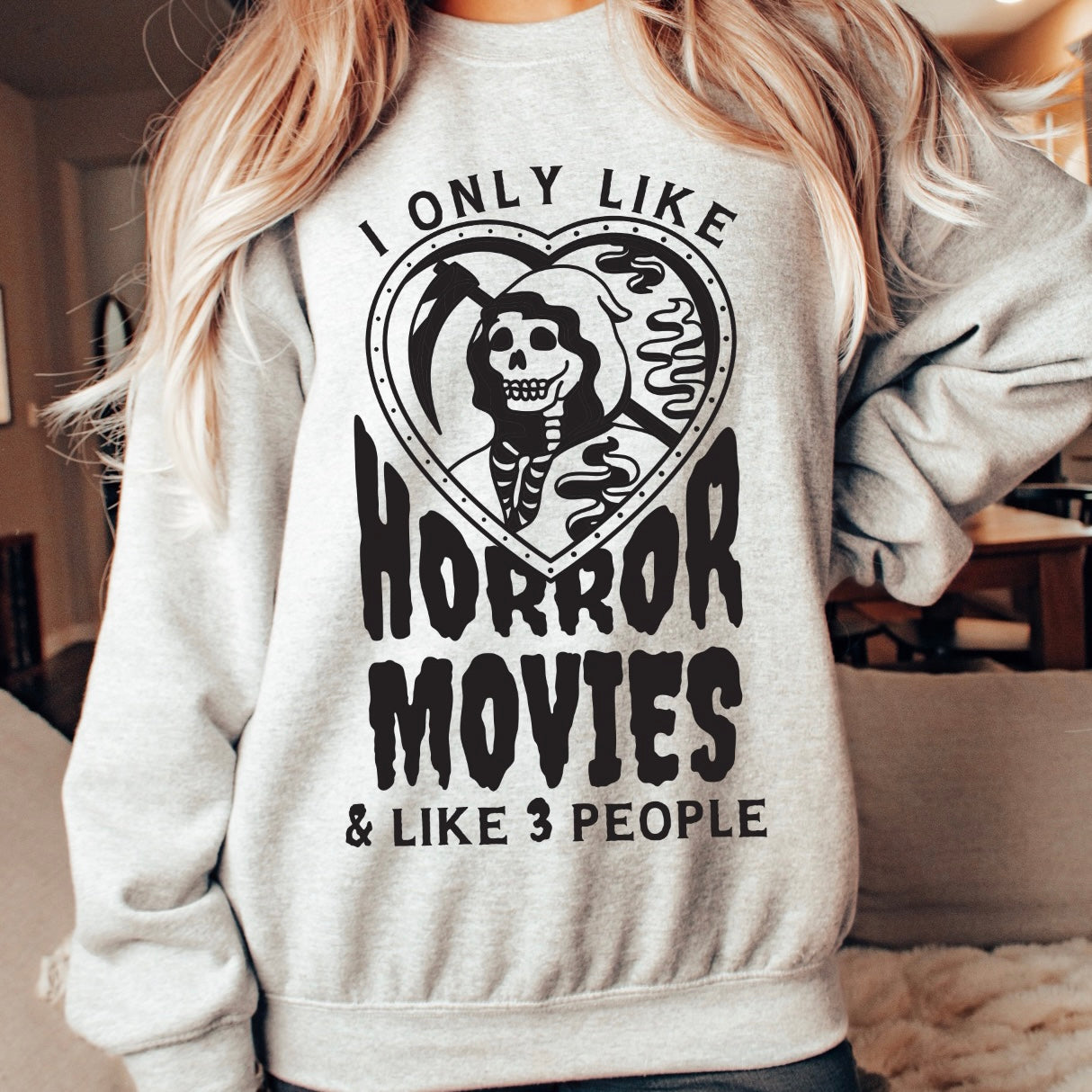 A person wears the Horror Movies • Ash Gray Pullover, a classic fit cotton-poly sweatshirt with a heart-shaped skeleton and the text: I only like horror movies & like 3 people, in a cozy, softly lit room.